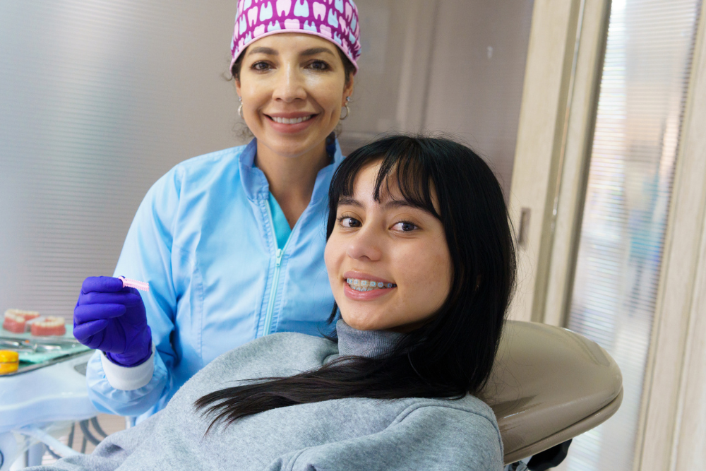 A dentist in blue scrubs and a patterned cap stands smiling beside a seated young patient with braces, creating a friendly, relaxed atmosphere. OrthoSynetics Marketing
