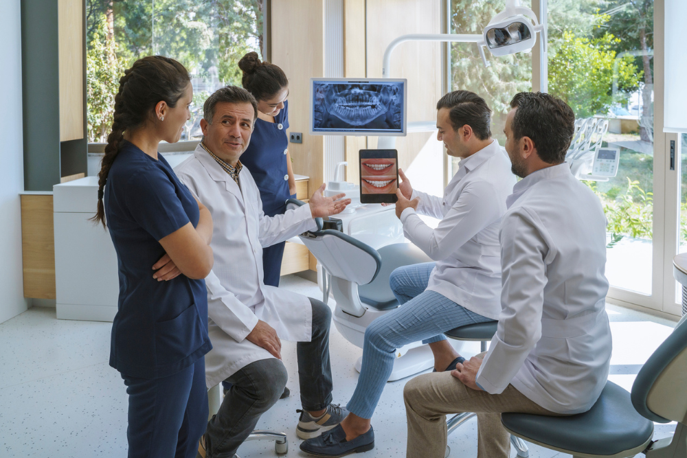 A group of five dental professionals in a clinic discusses a dental treatment plan. One holds a tablet showing a smiling mouth. The atmosphere is collaborative. Orthosynetics - Best Marketing Agency for Orthodontic Practices