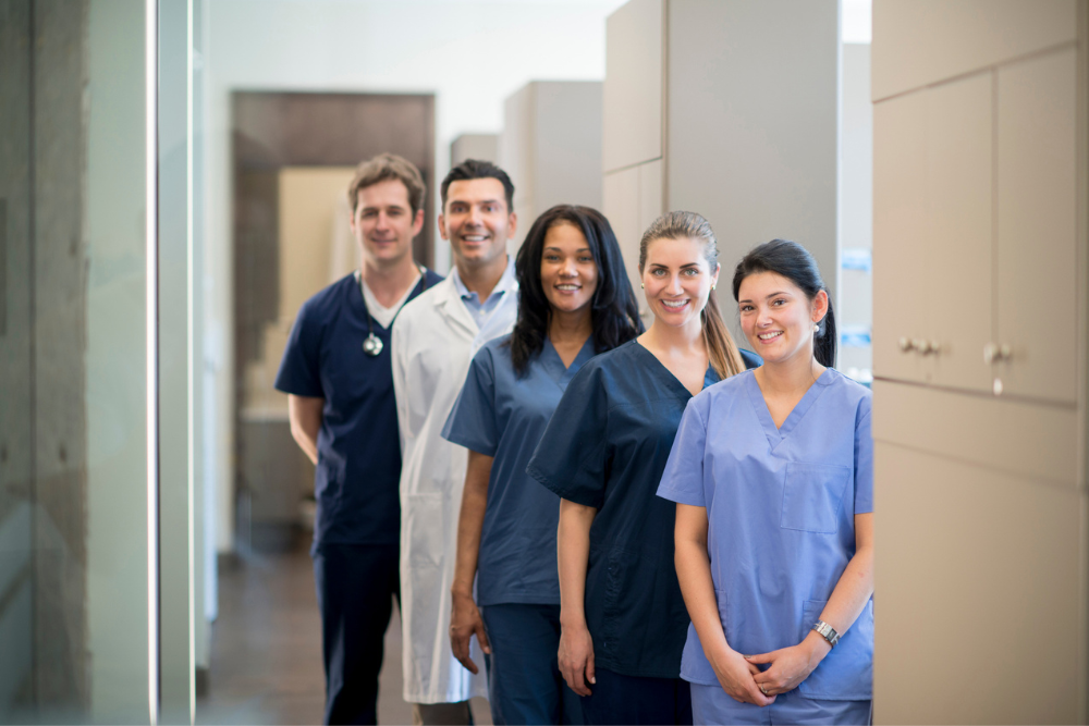 A diverse group of five healthcare professionals in scrubs and lab coats, smiling and standing in a well-lit hallway, conveying teamwork and positivity. Orthodontic & Dental Marketing FAQ