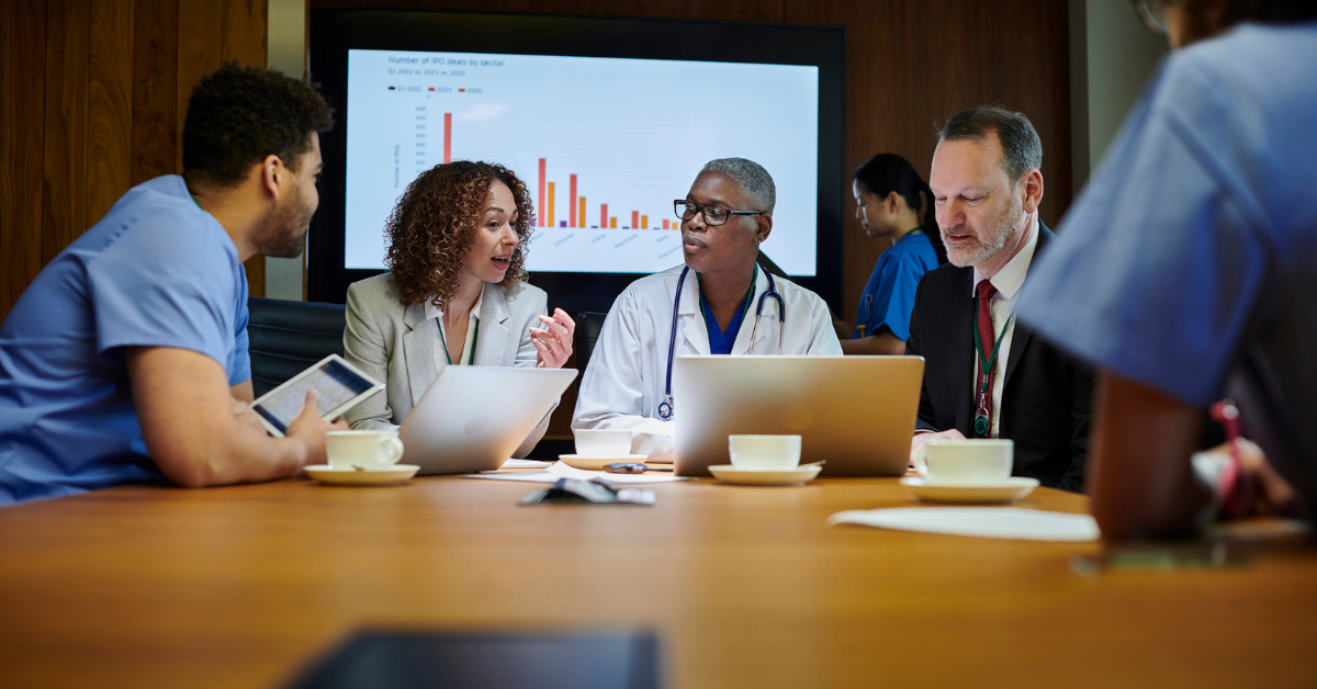 A diverse group of professionals in a meeting room, focused on laptops and discussing data displayed on a screen. The atmosphere is collaborative and serious.