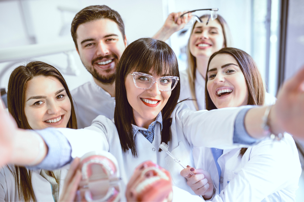 A group of five smiling dental professionals in white coats posing for a selfie. They hold dental tools and a model, conveying a cheerful, friendly atmosphere.