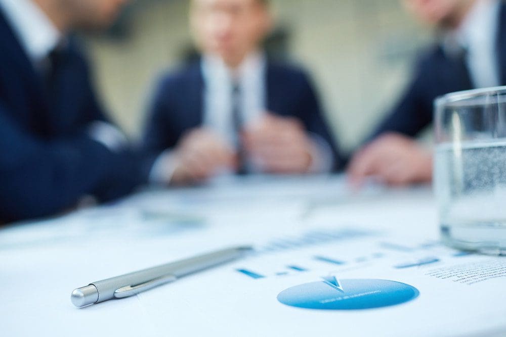 Image of business document and pen at workplace with group of colleagues interacting on background