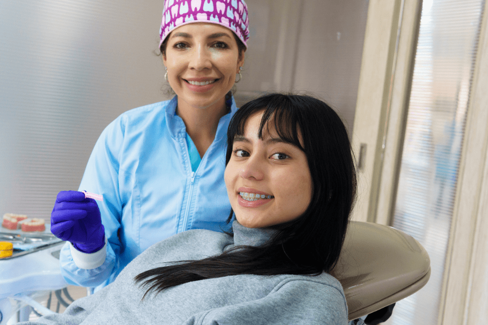 A dentist in blue scrubs and a patterned cap stands smiling beside a seated young patient with braces, creating a friendly, relaxed atmosphere. OrthoSynetics Marketing
