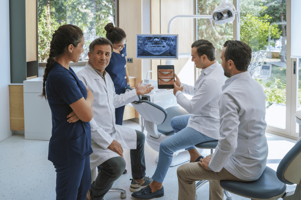 A group of five dental professionals in a clinic discusses a dental treatment plan. One holds a tablet showing a smiling mouth. The atmosphere is collaborative. Orthosynetics - Best Marketing Agency for Orthodontic Practices