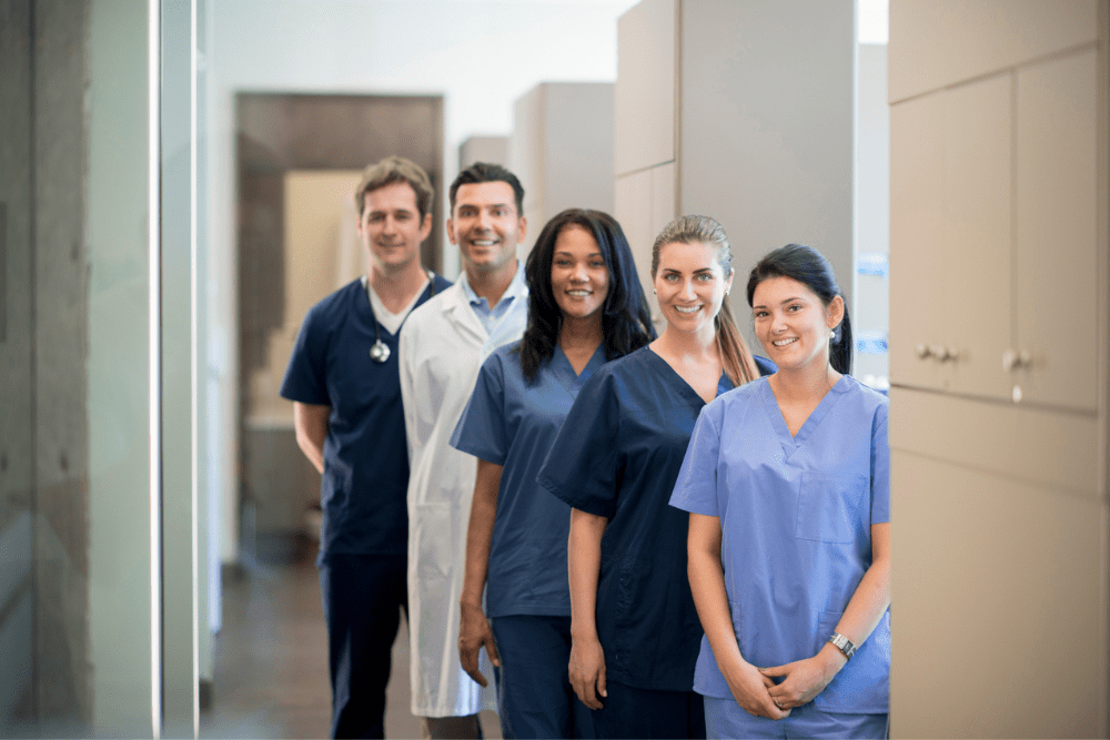 A diverse group of five healthcare professionals in scrubs and lab coats, smiling and standing in a well-lit hallway, conveying teamwork and positivity. Orthodontic & Dental Marketing FAQ